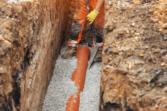 Ouvrier de construction installant un tuyau de drainage dans une tranchée à la lumière du jour, entouré de gravier et de terre