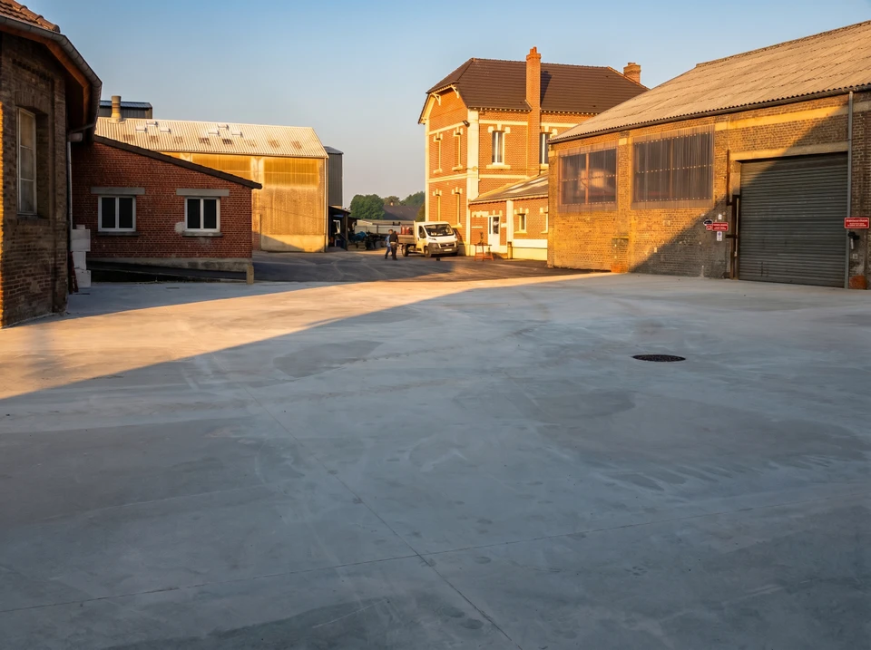 Chantier extérieur avec ouvrier en salopette bleue lissant une dalle de béton fraîchement coulée à l'aide d'une taloche